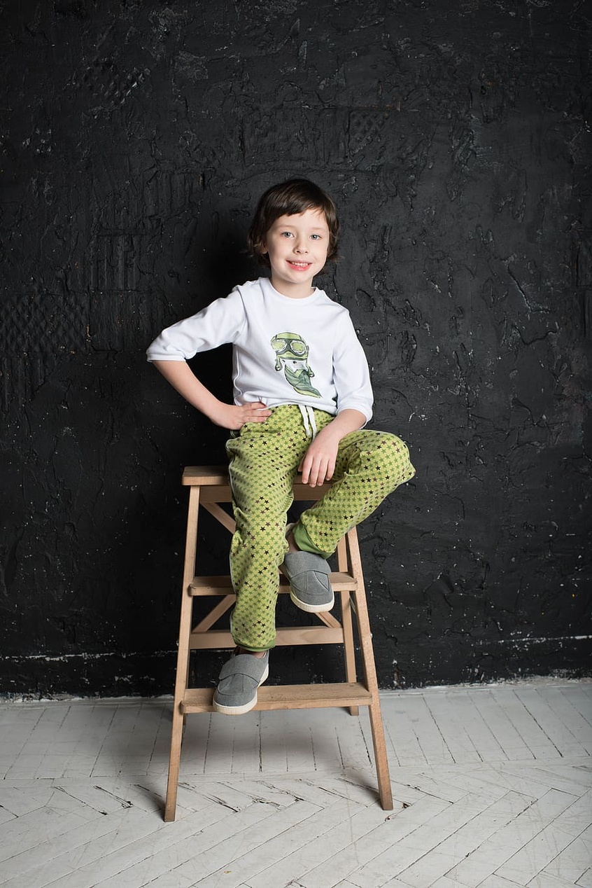 Ein kleiner Junge in einem modernen, schicken Outfit mit einem weißen Dinosaurier-Shirt und grüner Hose sitzt auf einem Hocker.